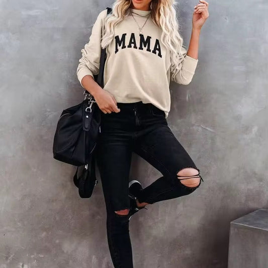 A woman wearing a beige sweatshirt with the word 'MAMA' printed on it, paired with black ripped jeans and black sneakers.