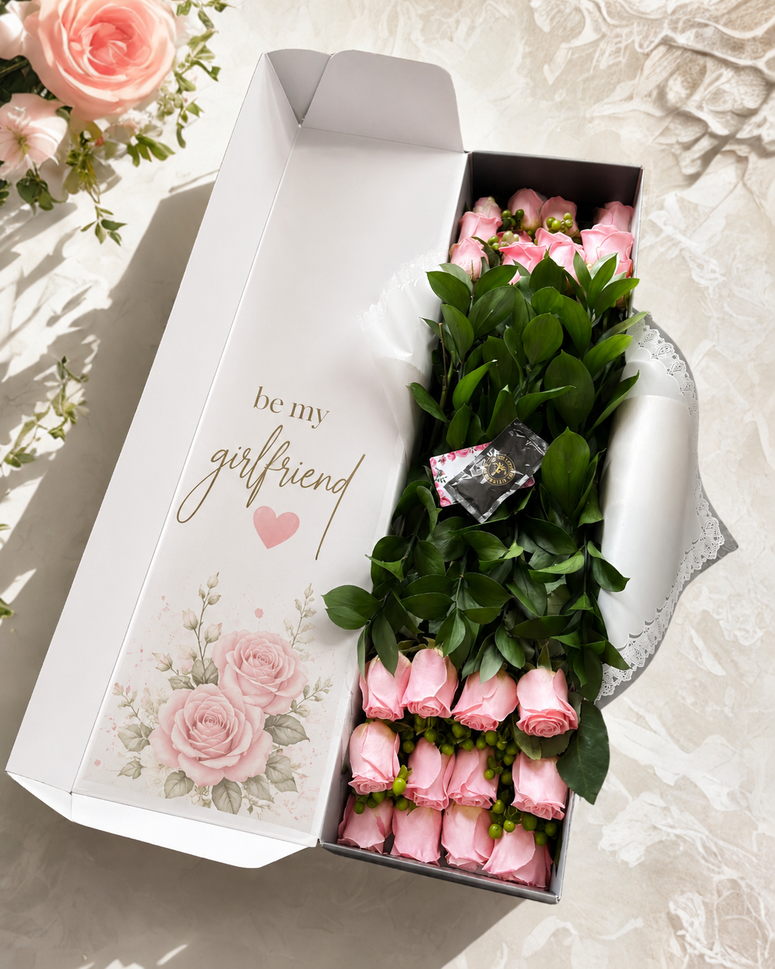 A gift box of pink roses with green leaves and a small chocolate, presented in an open white box printed with 'be my girlfriend' and rose illustrations, on a light marble surface.