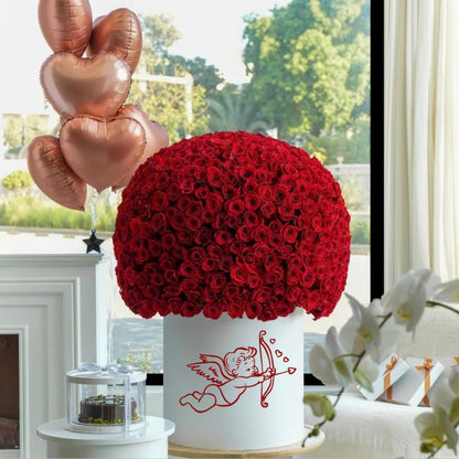Red rose arrangement in a white container with cupid design, heart-shaped balloons in the background.