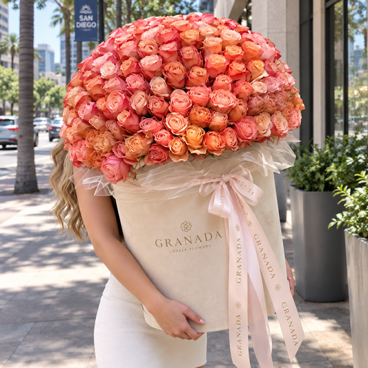 A woman on a city sidewalk holds a massive flower box overflowing with peach, coral, and orange roses. The box is cream-colored with a pale pink ribbon printed with the brand name GRANADA.