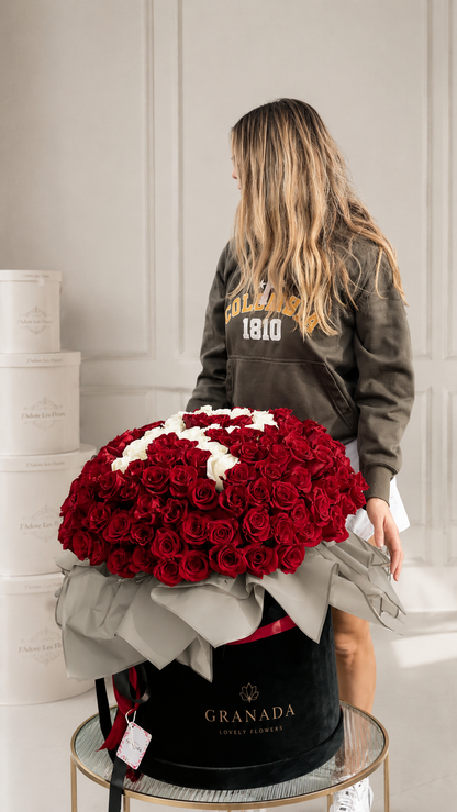 A woman in a dark olive hoodie stands beside a large round flower box filled with deep red roses and white rose accents, with the brand name GRANADA visible on the black container.