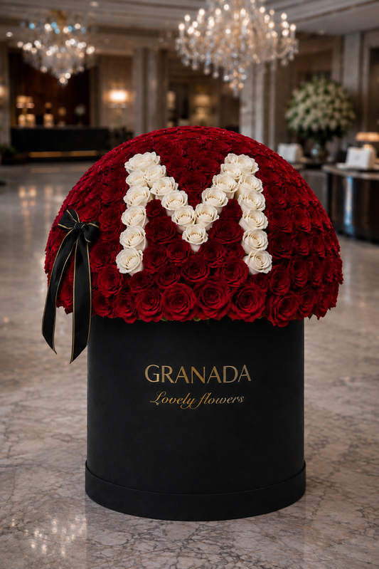 A large round floral arrangement of deep red roses with a white rose letter 'M' on top, presented in a black cylindrical box with gold 'GRANADA Lovely flowers' branding, set in a luxurious hotel lobby.