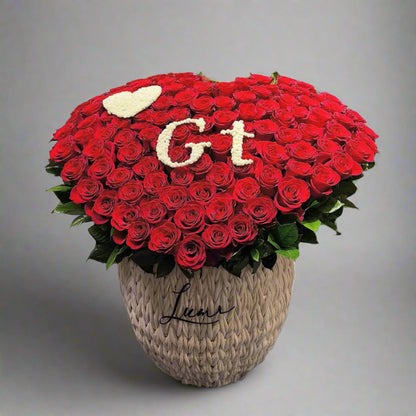 Heart-shaped arrangement of red roses with 'GT' and a heart on top, placed in a textured pot against a dark background.