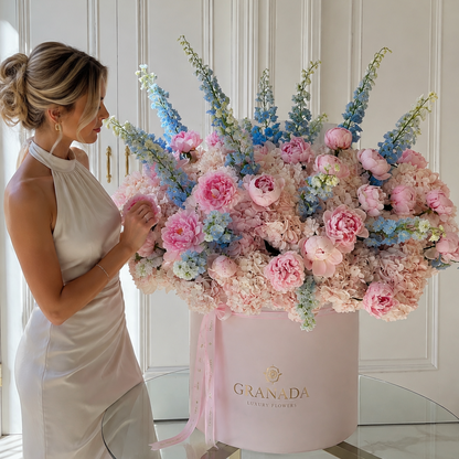 A woman in a sleeveless cream dress stands beside a large floral arrangement in a round pink box labeled GRANADA, placed on a glass-topped table in a bright indoor setting with white paneled doors behind.