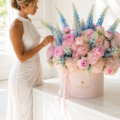A woman in a white sleeveless dress stands beside a large pastel flower arrangement in a round pink box labeled GRANADA, placed on a white table in a bright indoor setting.