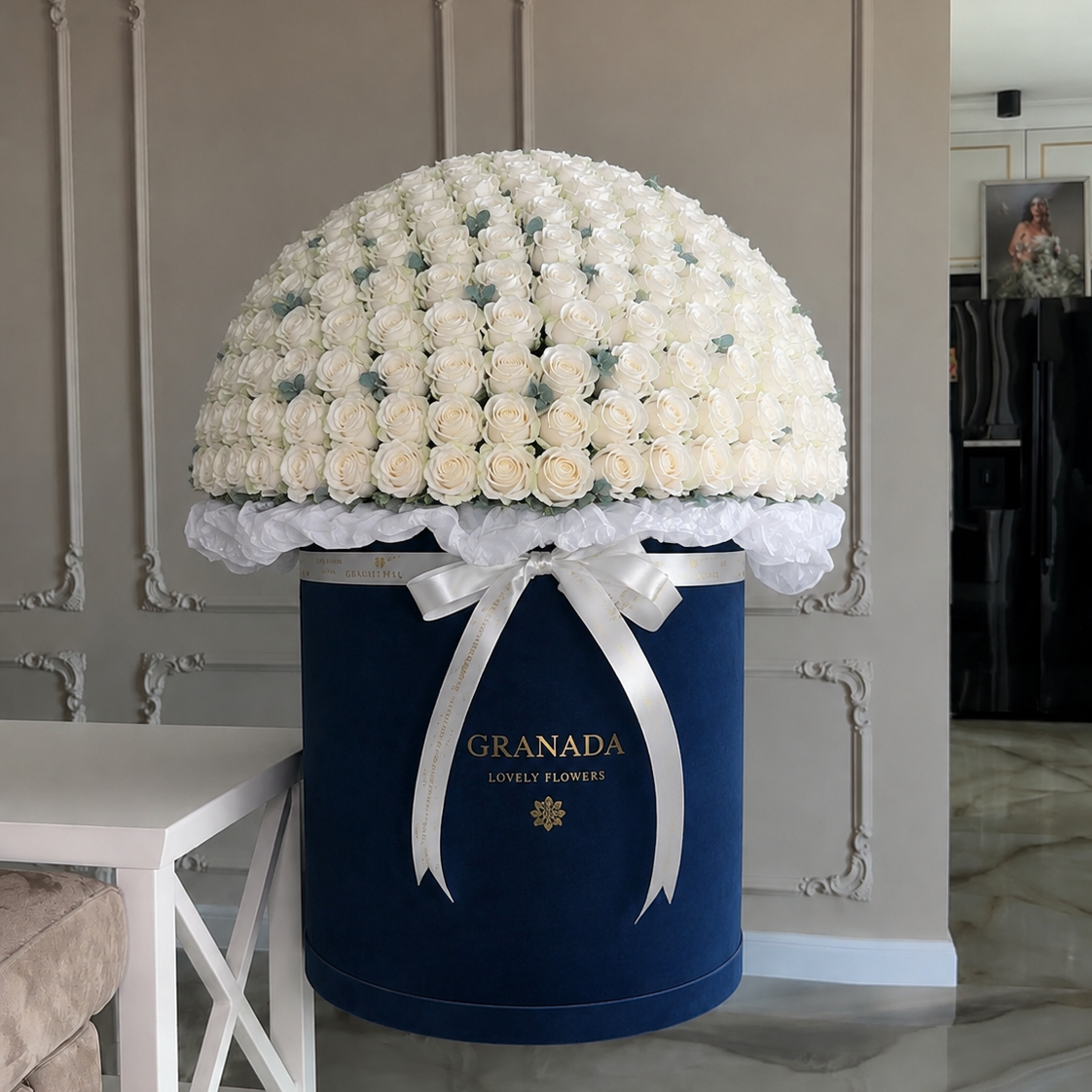 A large floral arrangement of white roses displayed on top of a navy blue cylindrical box with a white ribbon bow. The box is branded 'GRANADA JEWELRY HARVEST' in gold lettering, set against an elegant beige paneled wall.