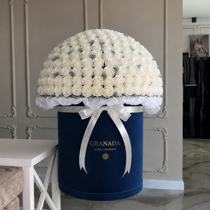 A large floral arrangement of white roses displayed on top of a navy blue cylindrical box with a white ribbon bow. The box is branded 'GRANADA JEWELRY HARVEST' in gold lettering, set against an elegant beige paneled wall.