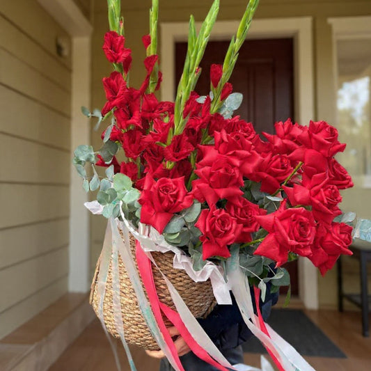 Bouquet of red flowers with green stems in a woven basket on a wooden floor.