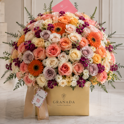 A large, vibrant floral arrangement in a square gold box branded GRANADA COFFEE FLORIST. The bouquet is a dense mix of orange, pink, peach, cream, and purple flowers with green foliage, set on a light marble surface against a neutral cream paneled wall.