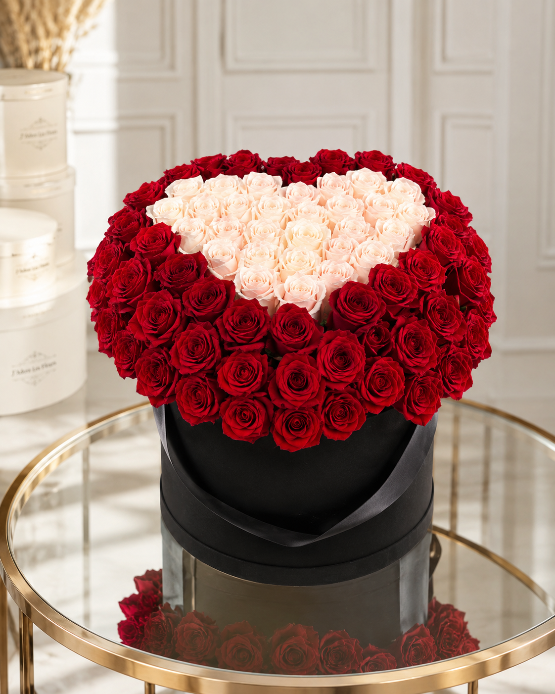 A heart-shaped rose arrangement in a black cylindrical box, featuring deep red roses around the outer edge and a pale blush pink heart in the center, displayed on a glass-topped table.