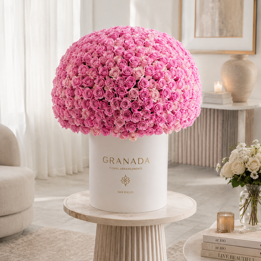 A large spherical bouquet of pink roses arranged in a white cylindrical box printed with the brand name GRANADA, displayed on a round pedestal in a softly lit, elegant interior setting.