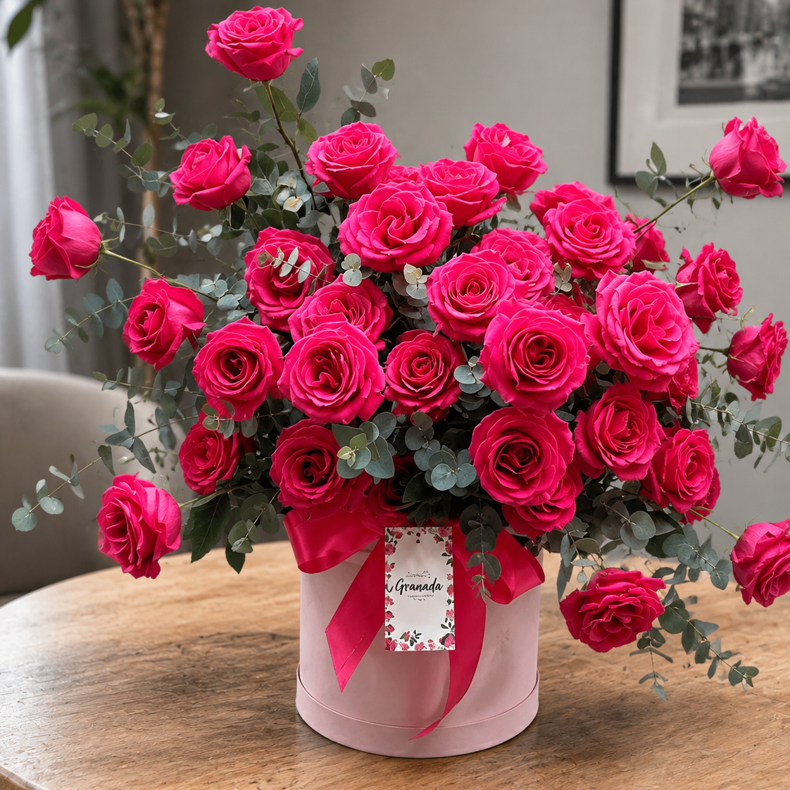A large bouquet of vibrant pink roses with green eucalyptus stems, presented in a light pink round box tied with a pink ribbon and a small tag that reads 'Grande'. The box sits on a round wooden table in a softly lit indoor setting.