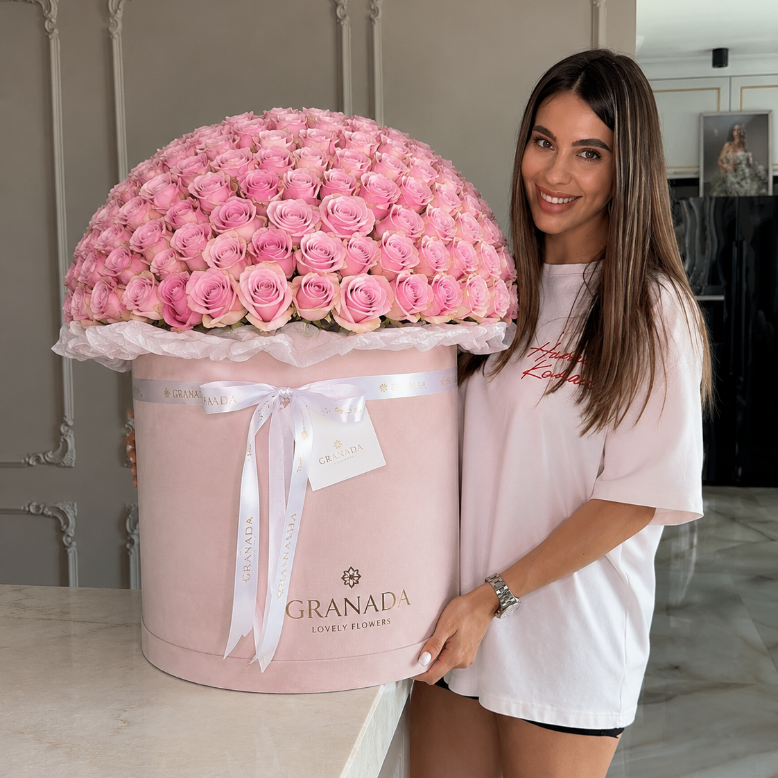 A smiling woman stands indoors beside a large round pink flower box filled with pink roses. The box is tied with a white ribbon and printed with the brand name GRANADA.