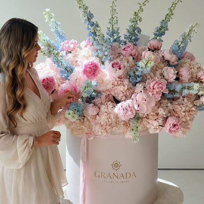 A woman in a light beige dress stands beside a large round pink box filled with a voluminous floral arrangement in pastel pink, peach, and blue, on a white pedestal in a bright studio setting. The box is branded GRANADA.