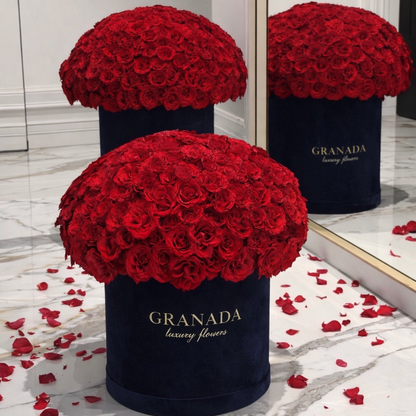Two black containers filled with red roses on a marble floor with scattered rose petals, branded 'Granada'.