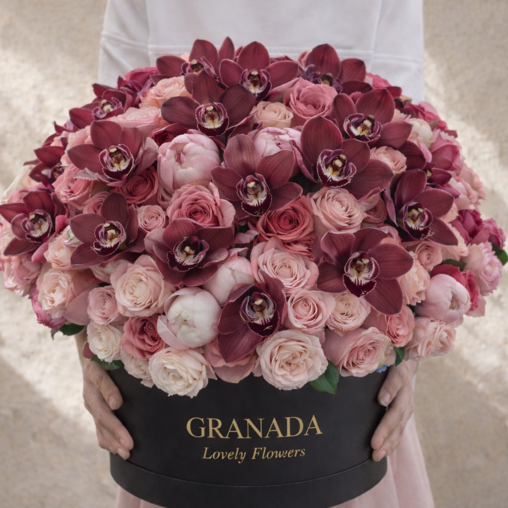 Person holding a bouquet of pink and purple flowers in a black box labeled 'Granada Lovely Flowers' on a beige background.