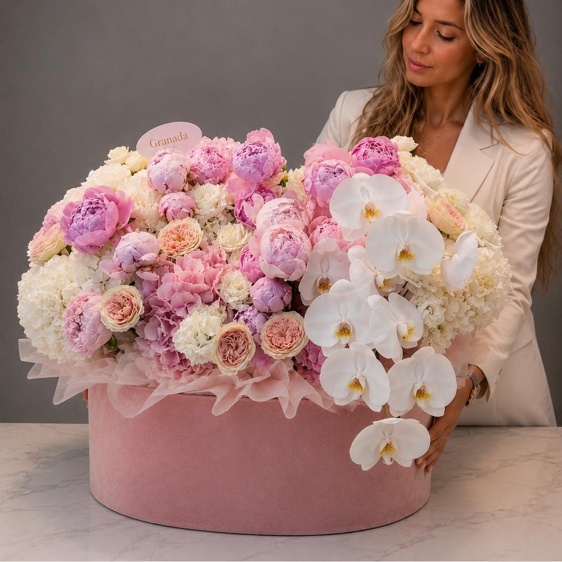 A woman in a white blazer stands beside a large round pink box filled with an abundant bouquet of pink, cream, and white flowers, including peonies and white orchids, against a gray studio background.